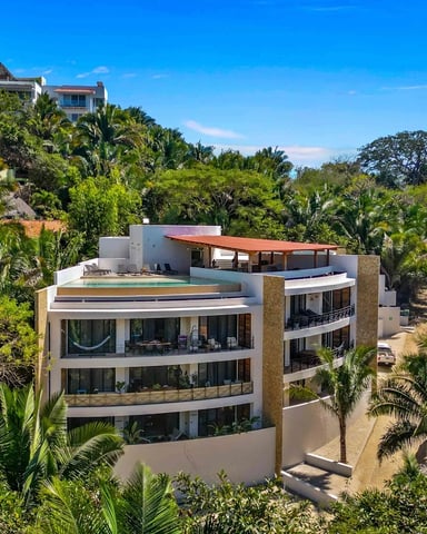 Modern multi-level villa with rooftop pool surrounded by tropical vegetation and palm trees on hillside.