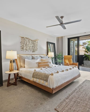 Spacious master bedroom with wood platform bed, cream bedding, ceiling fan, and sliding glass doors opening to tropical patio with palm trees