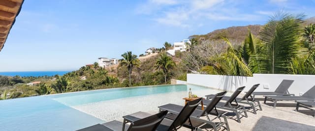 Infinity pool with black lounge chairs overlooking tropical hillside and ocean coastline