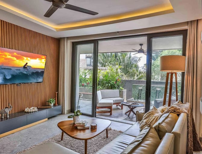 Modern living room with leather sofa, wooden coffee table, and sliding glass doors opening to tropical garden terrace with lounge seating